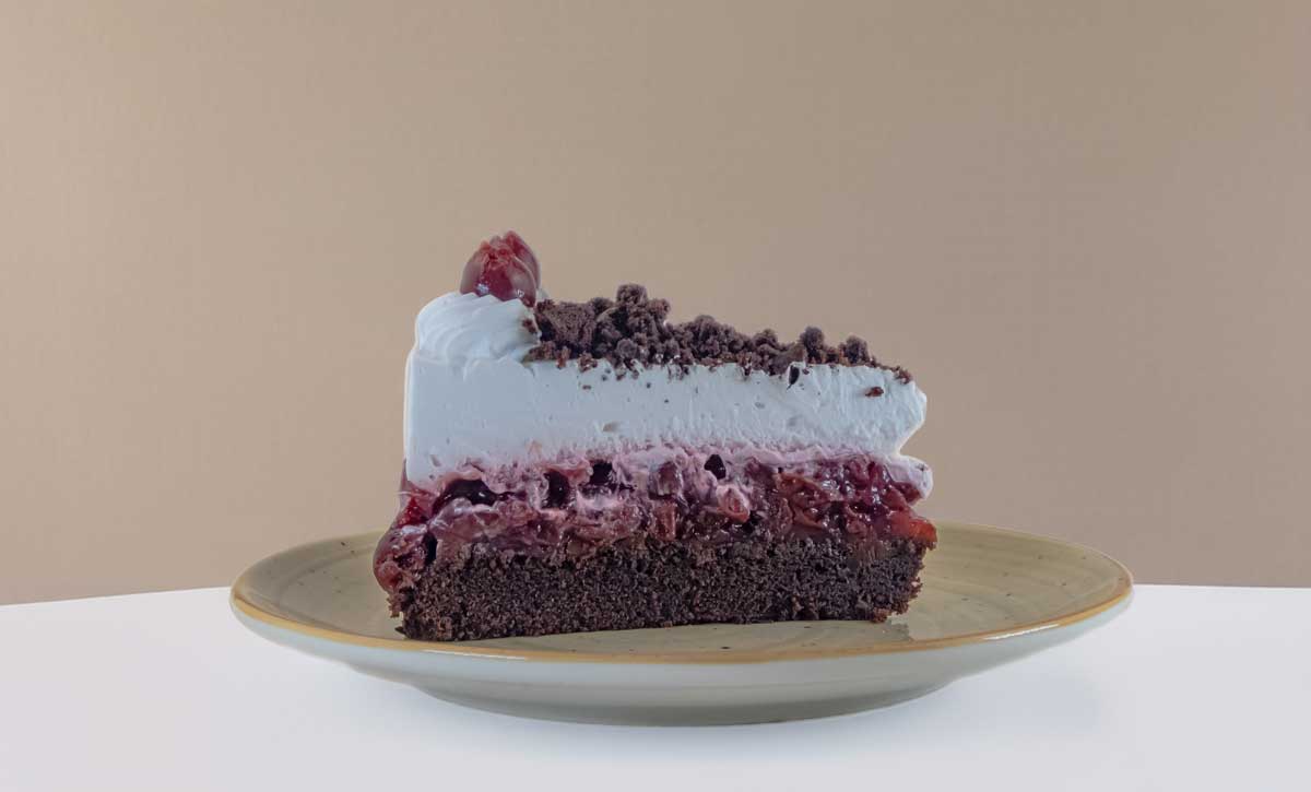 ein Stueck Schwarzwaelder Kirschtorte mit Schokoladenboden, Kirschfuellung und einer Schicht Schlagsahne mit Schokopulver bestreut