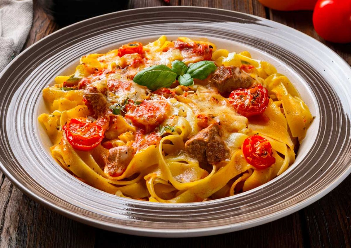 Teller mit Bandnudeln, Tomaten, Fleischstuecken mit Kaese gratiniert und Basilikum Deko